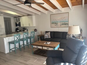 A modern kitchen with white cabinets, a dining area with teal chairs, a wooden coffee table, and a gray sofa. - SR-50 -1BR 1BA Courtyard Unit (Siesta Key)