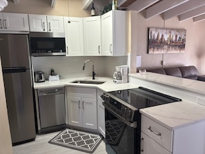 A compact kitchen with white cabinets, stainless steel appliances, a coffee maker, and a cityscape painting on the wall. - 1BR Siesta Key condo with king bed, sleep sofa, and courtyard views. (Siesta Key)