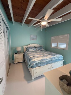 Ein Schlafzimmer mit Bett, Nachttisch, Lampe, Deckenventilator und einem Fenster mit Jalousien.