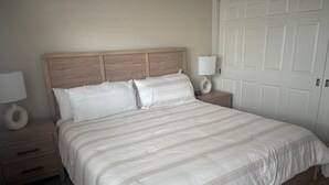 Een slaapkamer met een houten hoofdbord, een groot bed met witte beddengoed, twee nachtkastjes met lampen, en een witte deur op de achtergrond.