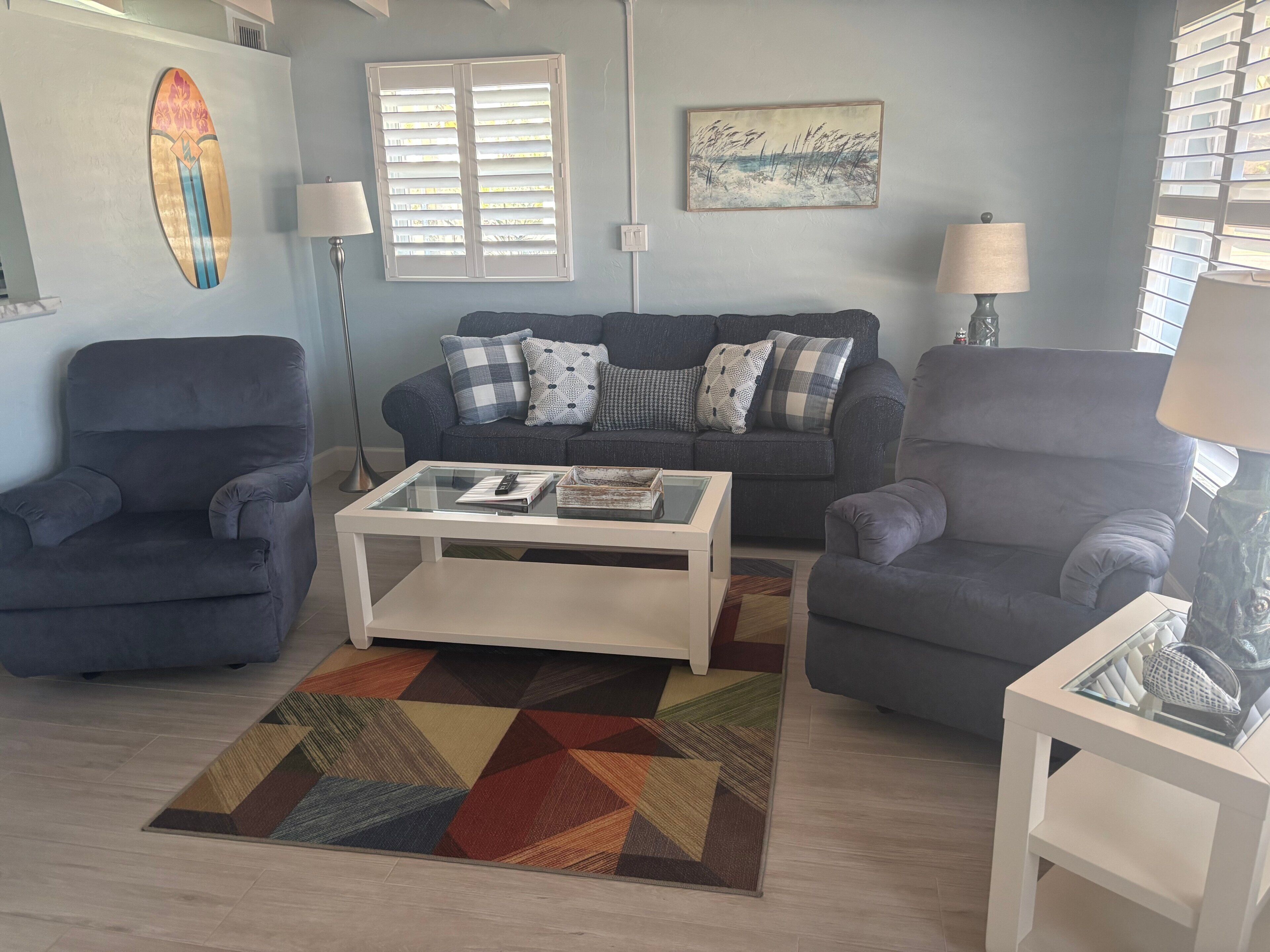 Un salon comprenant un canapé, deux fauteuils, une table basse, un tapis à motifs, une décoration sur le thème du surf et un tableau représentant une scène de plage.