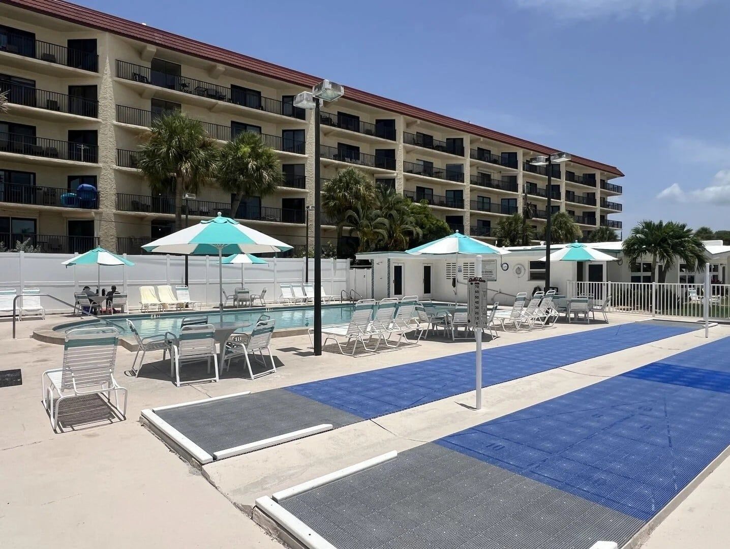 Une piscine d’hôtel entourée de chaises longues et de parasols, avec un bâtiment doté de nombreux balcons en arrière-plan.