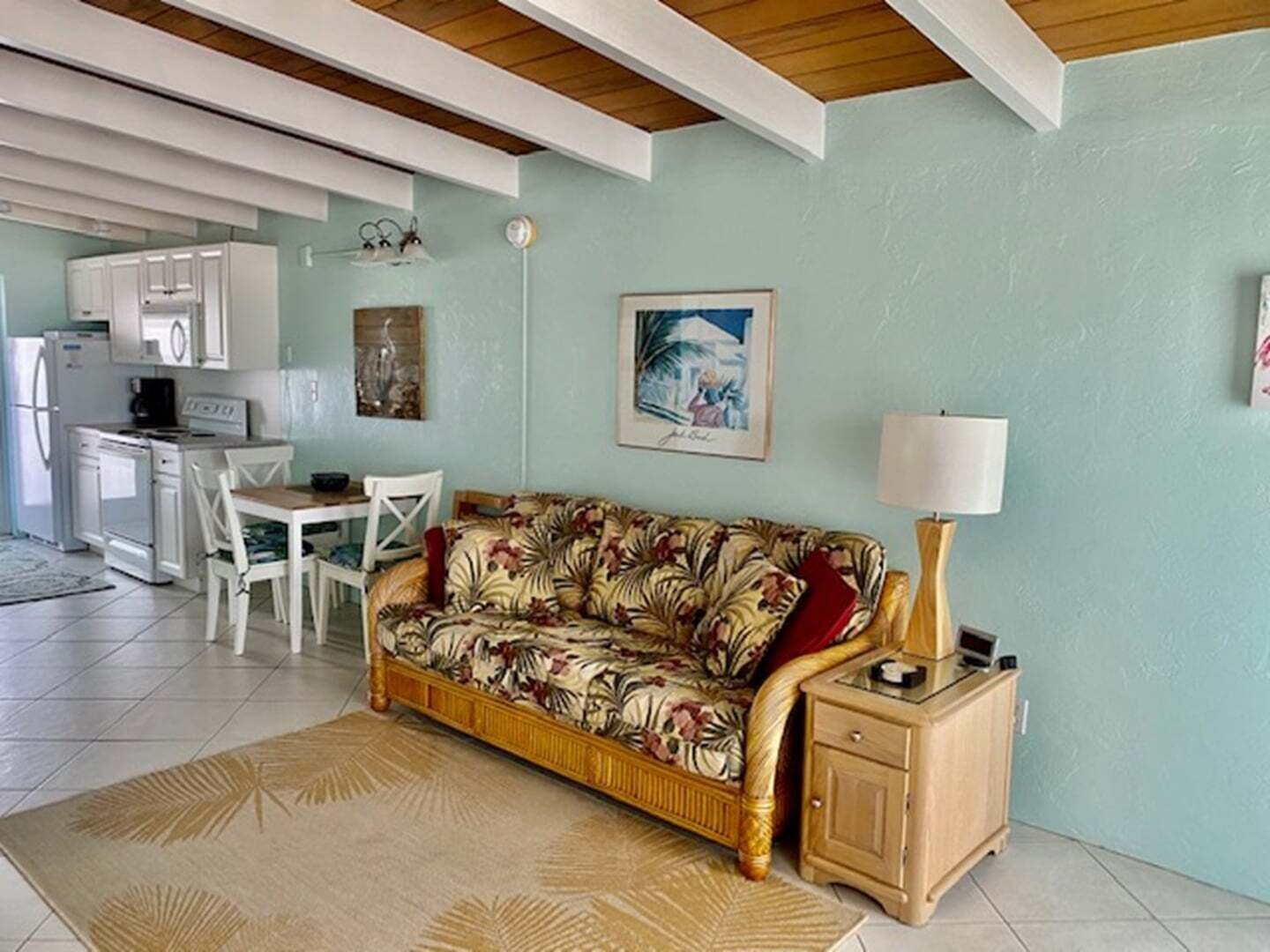 Una sala de estar con sofá, mesa de comedor, sillas, una cocina al fondo y una lámpara en una mesita de noche.
