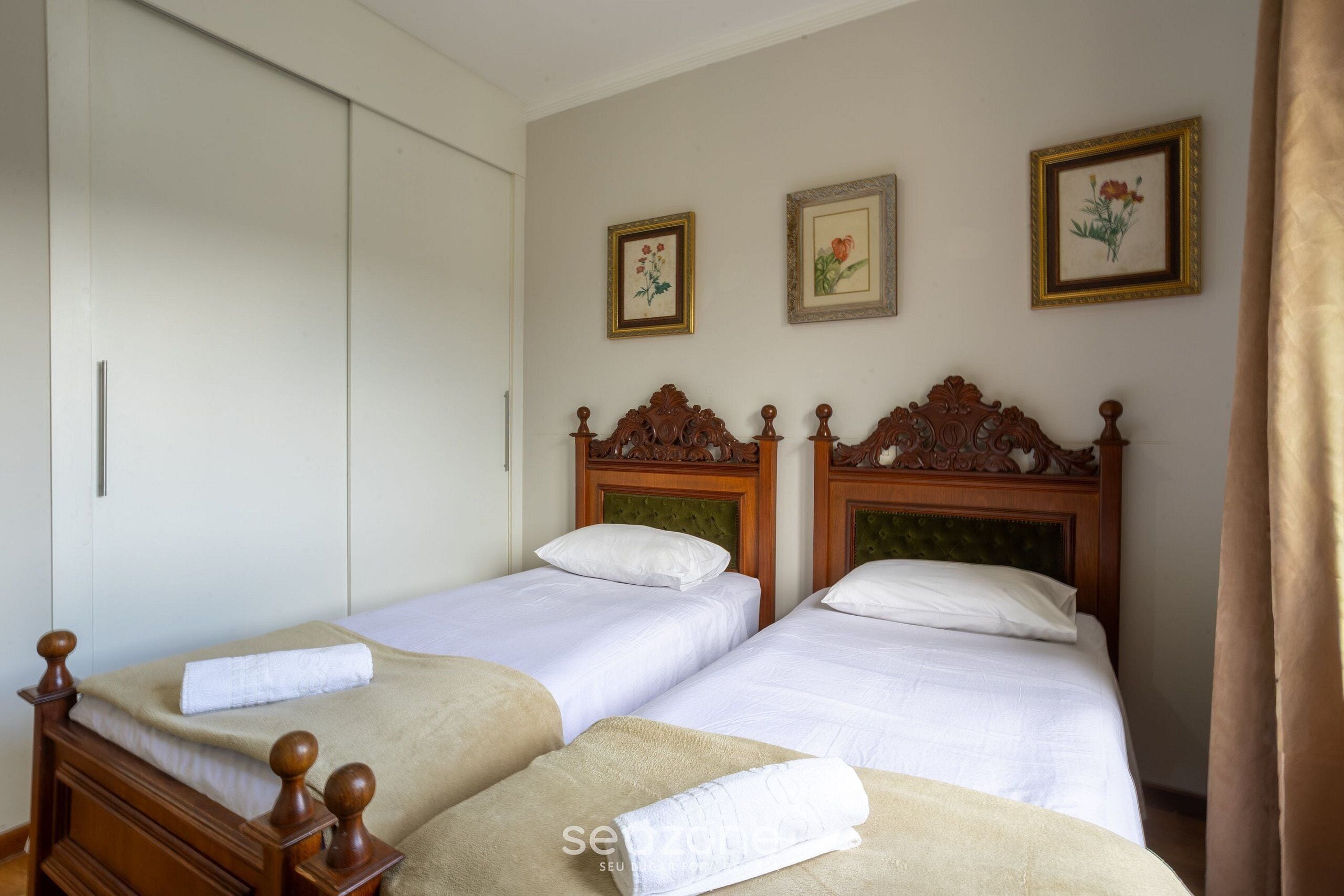 A room with two beds, a wardrobe, and three framed floral pictures on the wall.