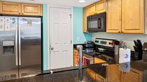 A kitchen with wooden cabinets, a stainless steel refrigerator, a microwave, a stove, and a water dispenser. - Pool & Palm Escape – Unit C208 (Biloxi)