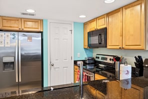 A kitchen with wooden cabinets, a stainless steel refrigerator, a microwave, a stove, and a water dispenser. - Pool & Palm Escape – Unit C208 (Biloxi)