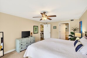 Ein Schlafzimmer mit einem Bett, einem Fernseher auf einer Kommode, einem Deckenventilator und einer Tür zu einem anderen Raum.