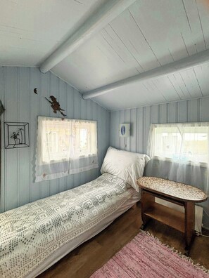 A small, single-bed room with a wooden bench, two windows with curtains, and a pink rug.