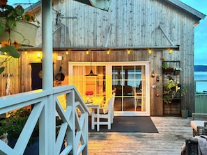 A wooden cabin with a deck, a dining area with chairs, and a view of the water.