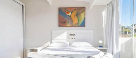 A modern bedroom with a large bed, a nightstand, a wall-mounted painting, and a view through a large window.