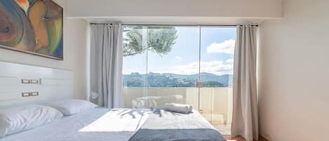 A bedroom with a bed, a painting, a view of the outdoors, and a window with curtains.