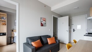 Ein modernes Wohnzimmer mit einem grauen Sofa, orangefarbenen Kissen, einem Esstisch mit gelben Stühlen und einem durch eine offene Tür einsehbaren Küchenbereich.