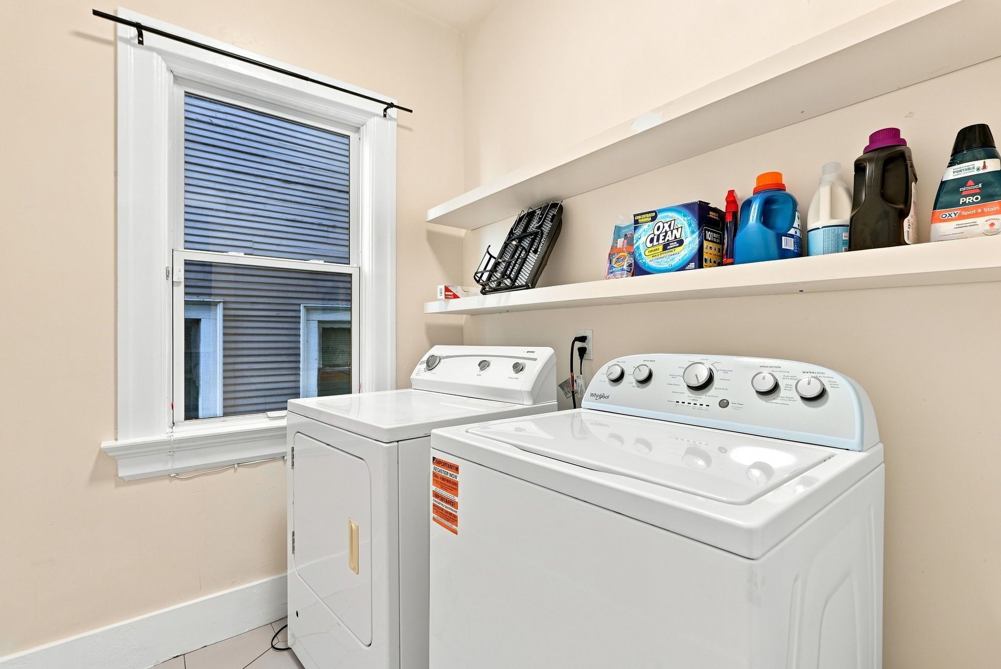 Une buanderie équipée de deux machines à laver et deux sèche-linge blancs, d’une fenêtre avec des stores et d’étagères contenant des produits de nettoyage.