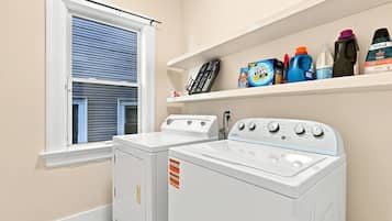 Une buanderie équipée de deux machines à laver et deux sèche-linge blancs, d’une fenêtre avec des stores et d’étagères contenant des produits de nettoyage.