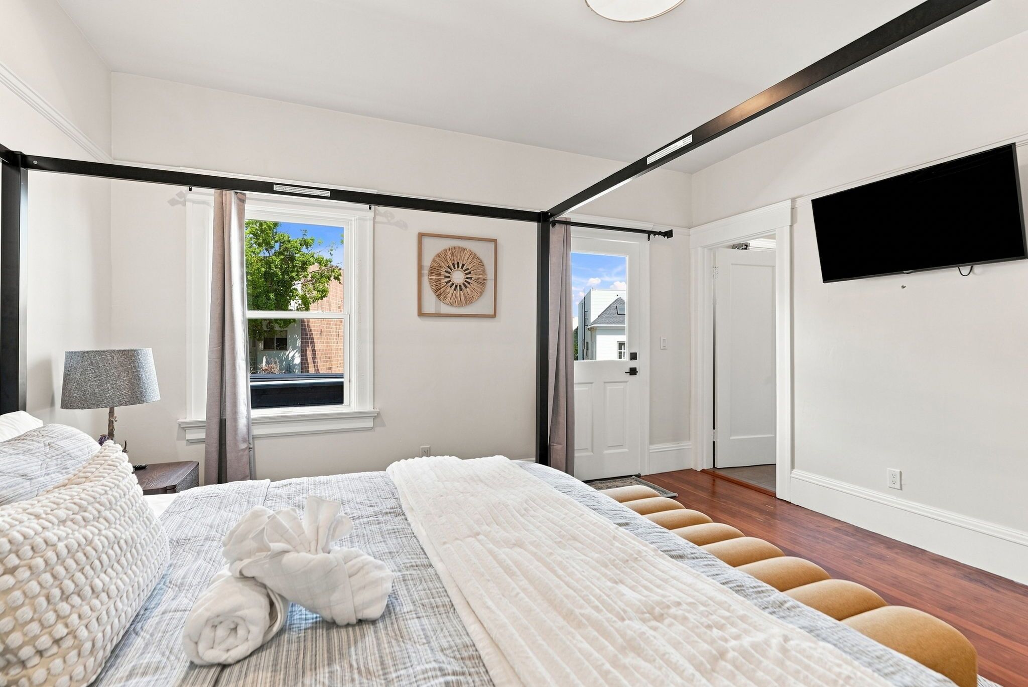 A hotel room with a large bed, a flat-screen TV, a window with a view of the outside, and a door leading to another room.