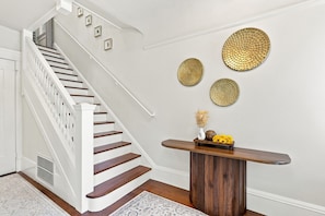 A staircase with white balusters and wooden steps, a white door, a radiator, a patterned rug, and a wooden console table with decorative items.