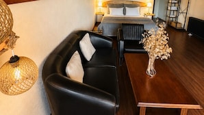 A hotel room with a wooden ceiling, a bed with a headboard, a black leather sofa, a wooden coffee table, a vase with dried flowers, and a painting on the wall.