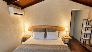 A bedroom with a sloped ceiling, a bed with a quilted bedspread, two bedside lamps, and a wall-mounted air conditioning unit.
