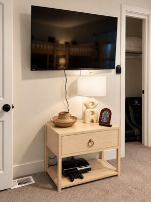 Une chambre avec un téléviseur fixé au mur, une table de chevet avec une lampe, un objet décoratif et une console de jeux.