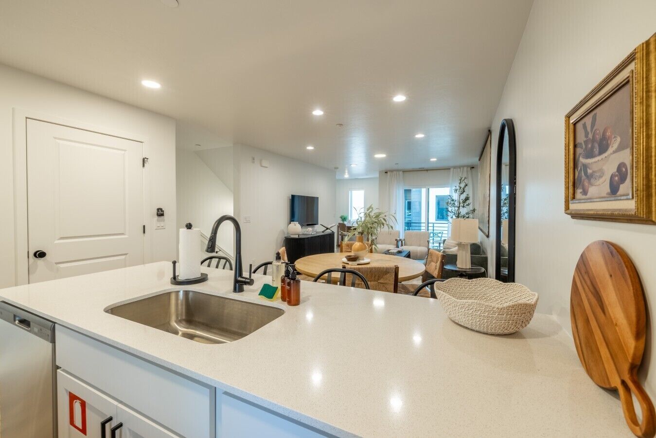 A modern kitchen with a double sink, a dining area with a round table, and a living space with a TV and plants.