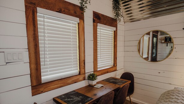 Une pièce avec un bureau en bois, deux fenêtres équipées de stores, un miroir rond et une petite plante posée sur le bureau.