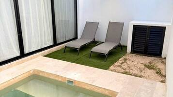 A modern outdoor pool area with a clear glass pool, two lounge chairs, and a pergola with string lights.