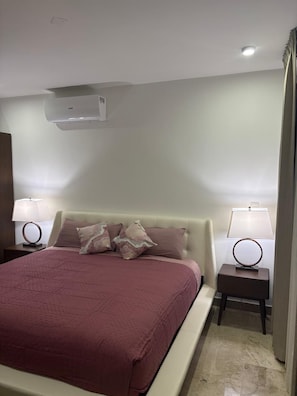 Habitación de hotel con una cama, dos mesitas de noche con lámparas, un equipo de aire acondicionado y una pared lisa.