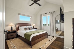Une chambre à coucher comprenant un lit, des tables de chevet, un ventilateur de plafond, une fenêtre avec des rideaux et un miroir.