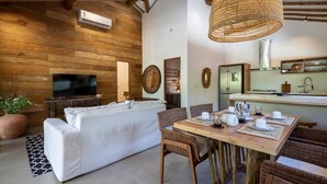 A modern living room with a white sofa, a dining table set for four, and a kitchen area with a refrigerator and a hanging light fixture. - Flamboyant Residence Quadrado (Porto Seguro)