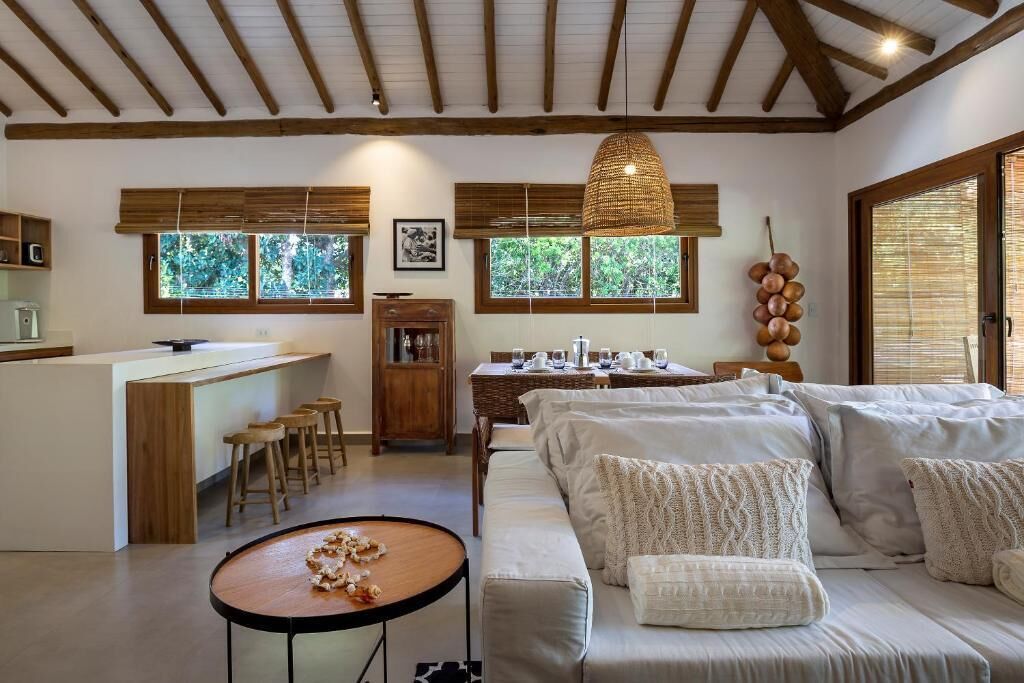 Un interior moderno con cocina, comedor y sala de estar. Se observan vigas de madera en el techo, ventanas amplias y una lámpara colgante.