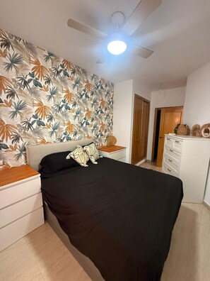 Een slaapkamer met een bed, nachtkastje, dressoir, plafondventilator en een muur met een bladpatroon.