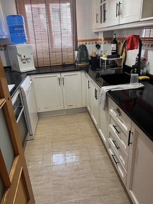 A kitchen with white cabinets, a black countertop, a water dispenser, and a window with blinds.