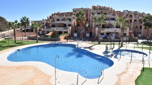 Ein Swimmingpool mit einer umlaufenden Terrasse, Palmen und einem mehrstöckigen Gebäude im Hintergrund.