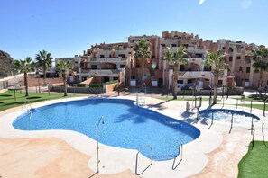 Ein Swimmingpool mit einer umlaufenden Terrasse, Palmen und einem mehrstöckigen Gebäude im Hintergrund.