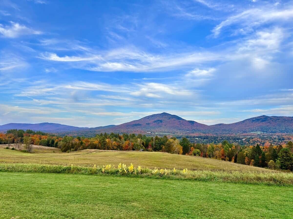 Un paisaje con una montaña, campos cubiertos de hierba y árboles con follaje otoñal.
