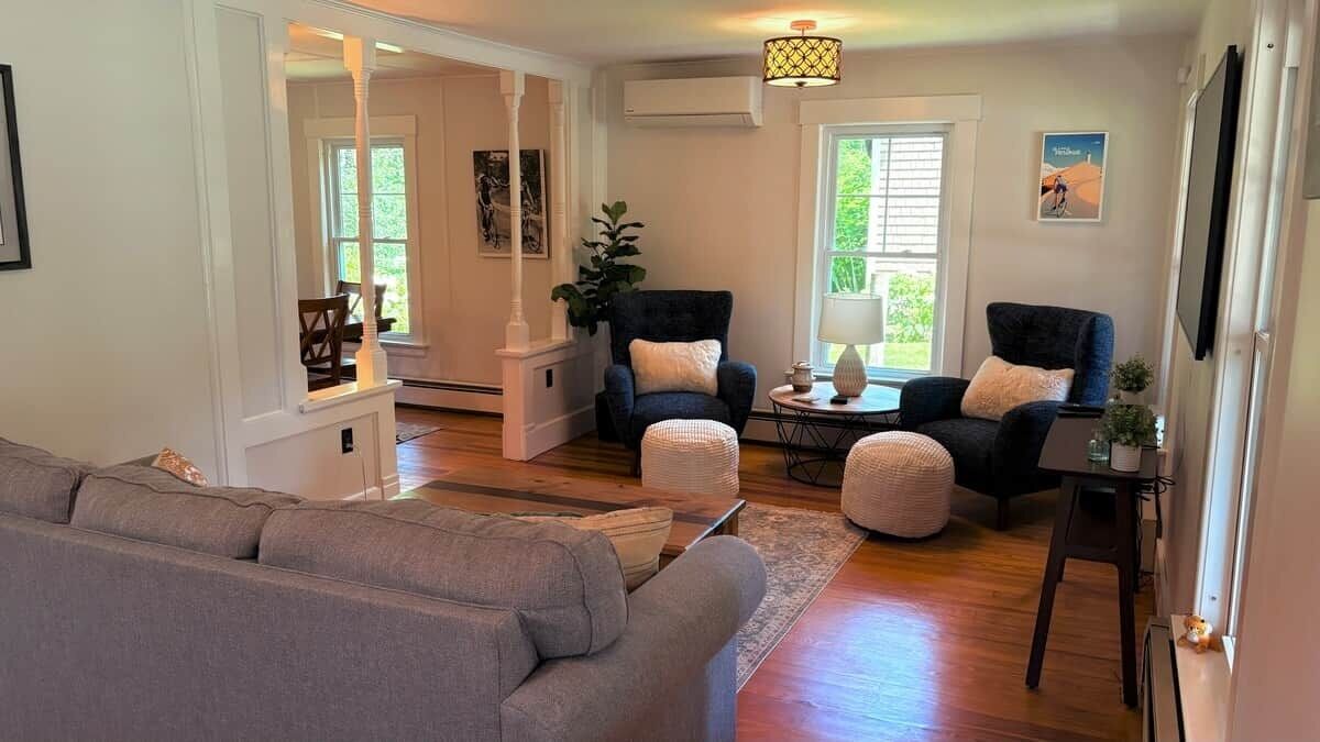 Un salon comprenant un canapé, deux fauteuils, une table basse et une lampe. Il y a une fenêtre avec des stores, un tableau au mur et une petite table sur laquelle est posée une plante.