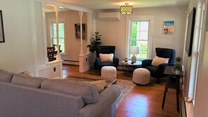 A living room with a sofa, two armchairs, a coffee table, and a lamp. There is a window with blinds, a picture on the wall, and a small table with a plant.