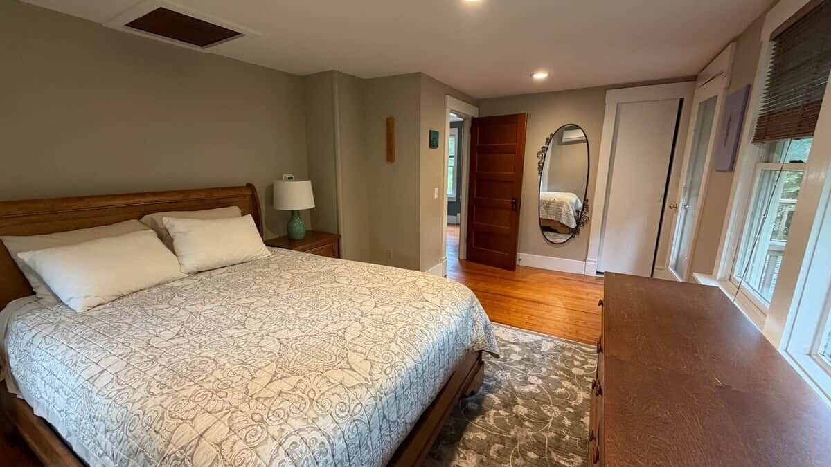 Ein Schlafzimmer mit Bett, Nachttisch, Lampe, Holzboden und Blick ins Freie.