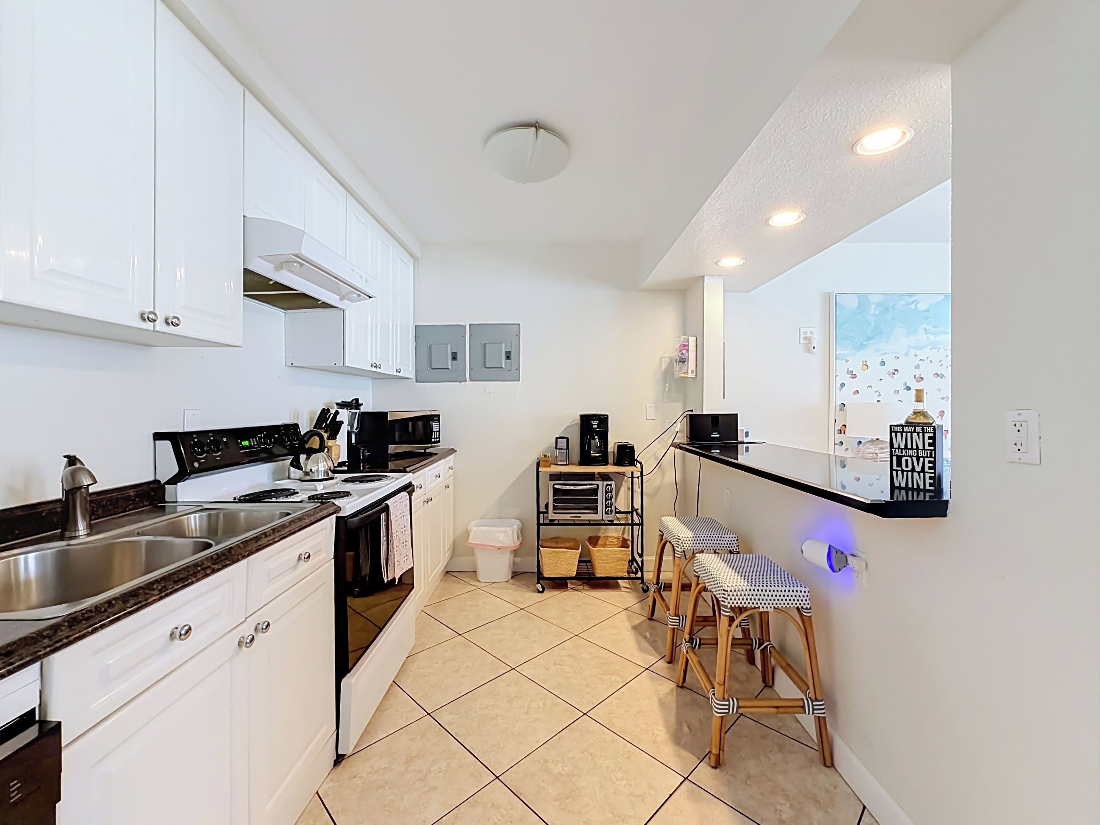 A modern kitchen with white cabinets, a stainless steel sink, a stove, and a coffee maker. There are two bar stools and a small table with a coffee machine.