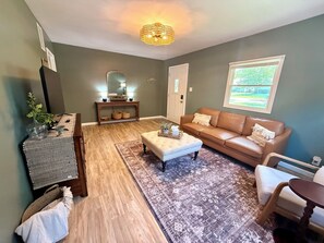 Ein Wohnzimmer mit einem braunen Ledersofa, einem weißen Hocker, einem Holzkonsole und einem gemusterten Teppich.