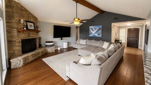 Ein Wohnzimmer mit Kamin aus Stein, einem großen Ecksofa, einem Flachbildfernseher und einem Deckenventilator.