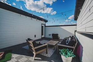 En udendørs terrasse med et træbord og stole, en potteplante og lyskæder.