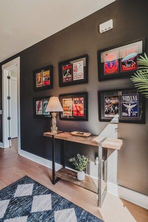 A hallway with framed concert posters on the wall, a wooden console table with a lamp, and a potted plant. - Nashville Legendz: Rooftop Deck (Nashville)