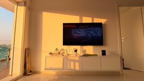 A modern living room with a large flat-screen TV mounted on the wall, a white entertainment unit below, and a sliding glass door leading to an outdoor area.