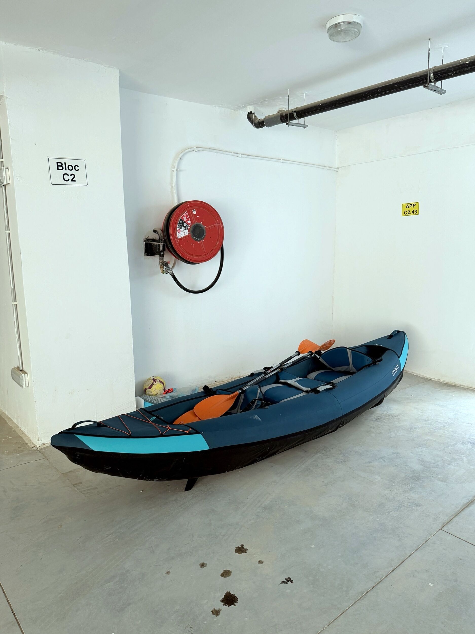 Un kayak en una habitación con paredes blancas, un extintor y una tubería en el techo.