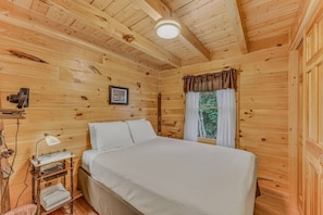 Een slaapkamer in een houten chalet, met een bed, nachtkastje, lamp en een raam met gordijnen.