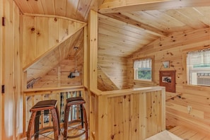 A wooden interior with a sloped ceiling, a window with a view, and two bar stools.