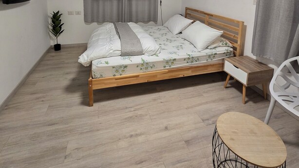 A bedroom with a bed, a bedside table, a chair, a small round table, a TV, a plant, and an air conditioning unit. - Queen 1 - Tranquil 1-bedroom hotel suites in Kiryat Ata (Kiryat Ata)