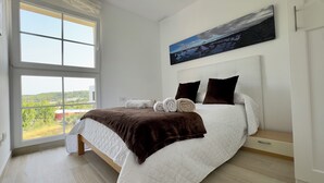 A bedroom with a large window, a bed with a quilted white duvet, two dark pillows, and a brown throw blanket.