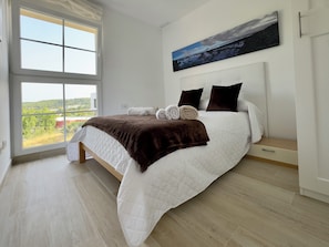 A bedroom with a large window, a bed with a quilted white duvet, two dark pillows, and a brown throw blanket.
